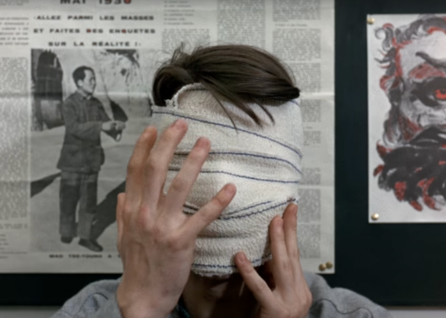 Still from Godard’s 1967 film, La Chinoise. The actor Jean-Pierre Léaud brings his hands up to his face, which is covered entirely by a white bandage.