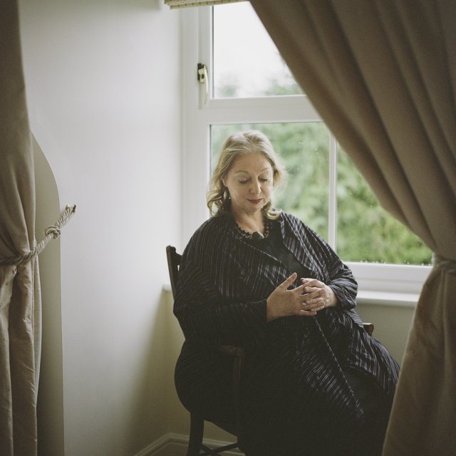 Portrait of Hilary Mantel in 2020. The author is seated by a window. She laces her hand in front of her stomach and is gazing downward. The light is soft.