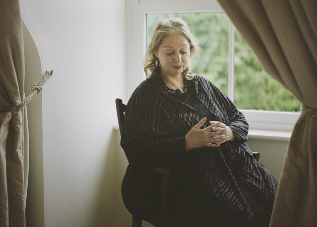 Portrait of Hilary Mantel in 2020. The author is seated by a window. She laces her hand in front of her stomach and is gazing downward. The light is soft.